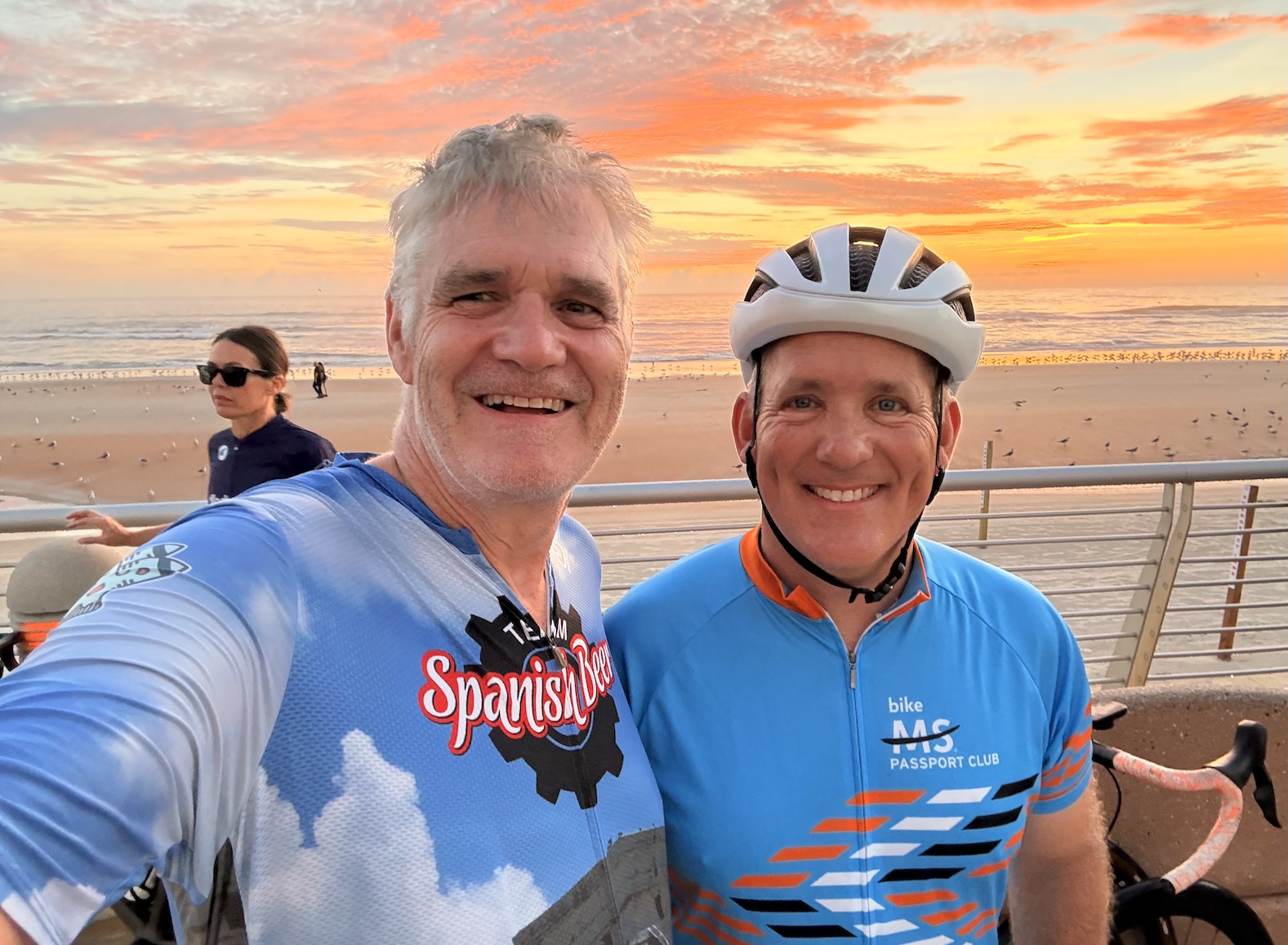 Casey in a Team Spanish Beer jersey and Curt in a Bike MS Passport Club jersey smiling on a beach boardwalk at sunrise