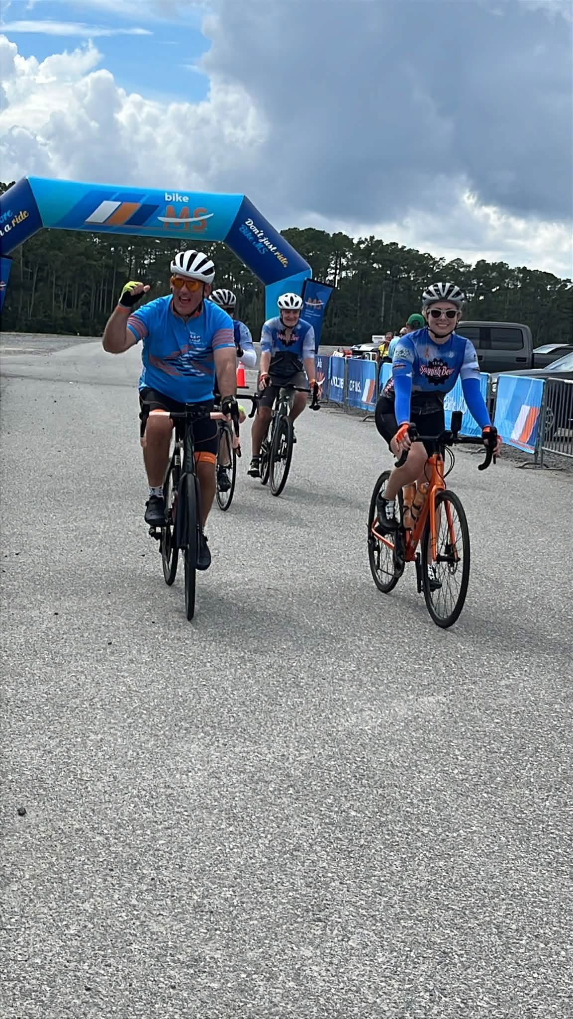 Curt pumping his fist and Selena smiling on her orange bike as they ride away from the Bike MS arch