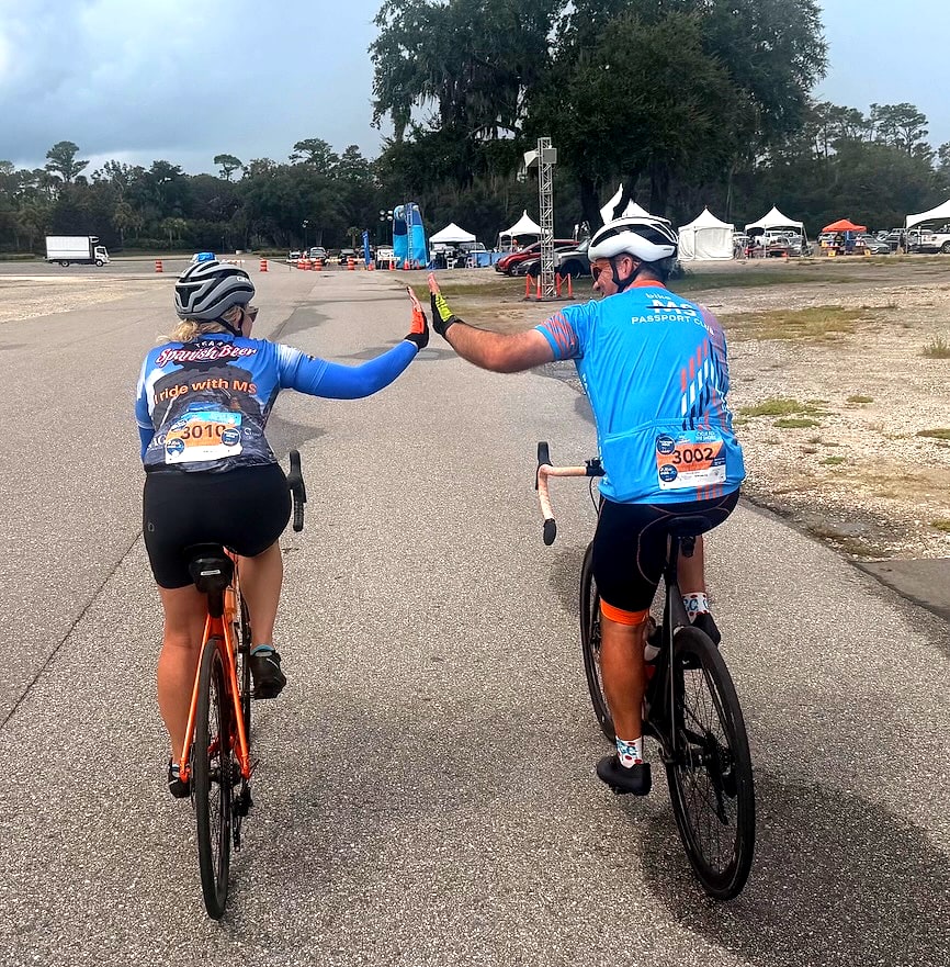 Selena in an I Ride with MS Team Spanish Beer jersey high-fiving Curt as they ride their bikes toward the finish