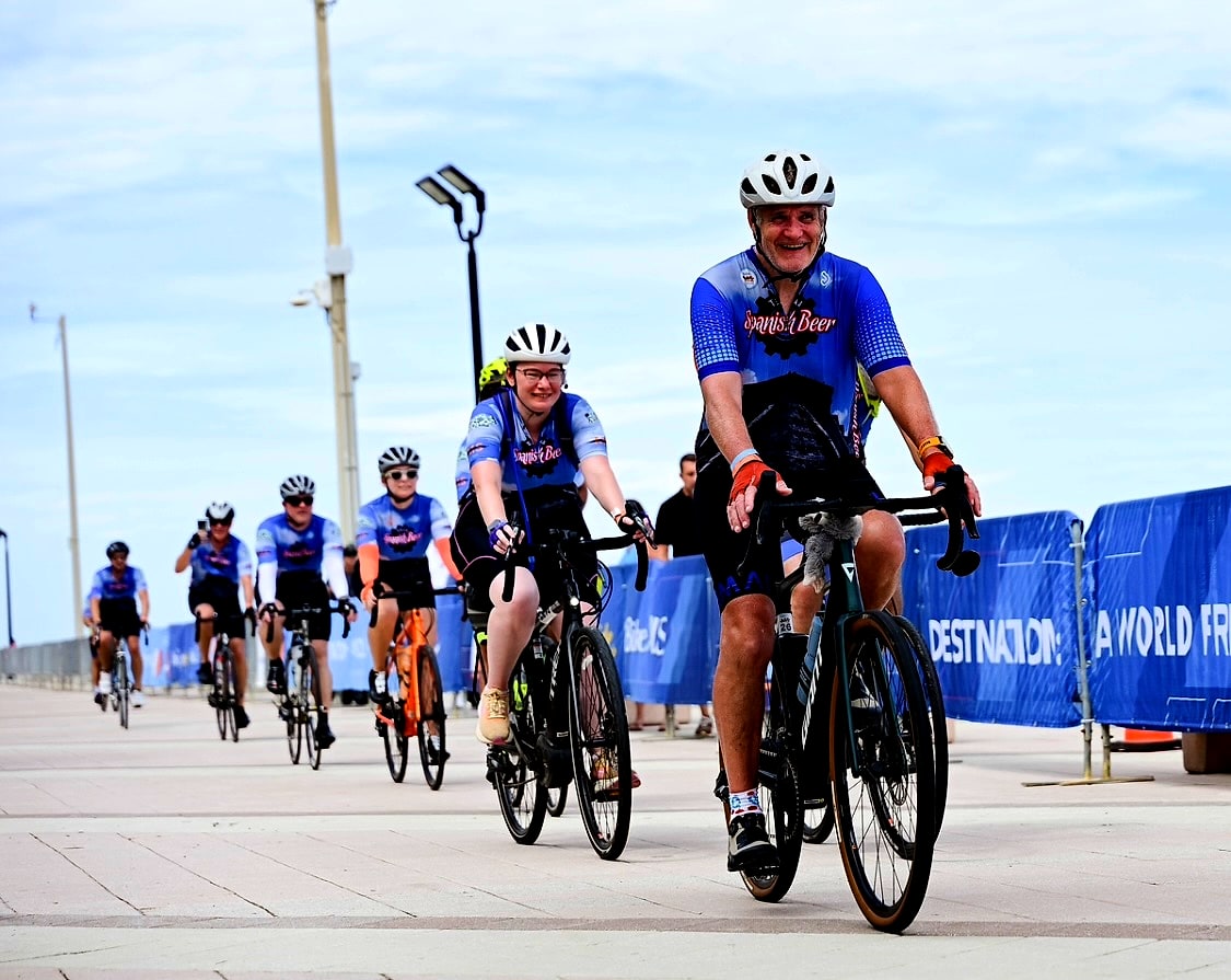 Team Spanish Beer riders in blue jerseys cycling along the beach boardwalk toward the Day 1 finish